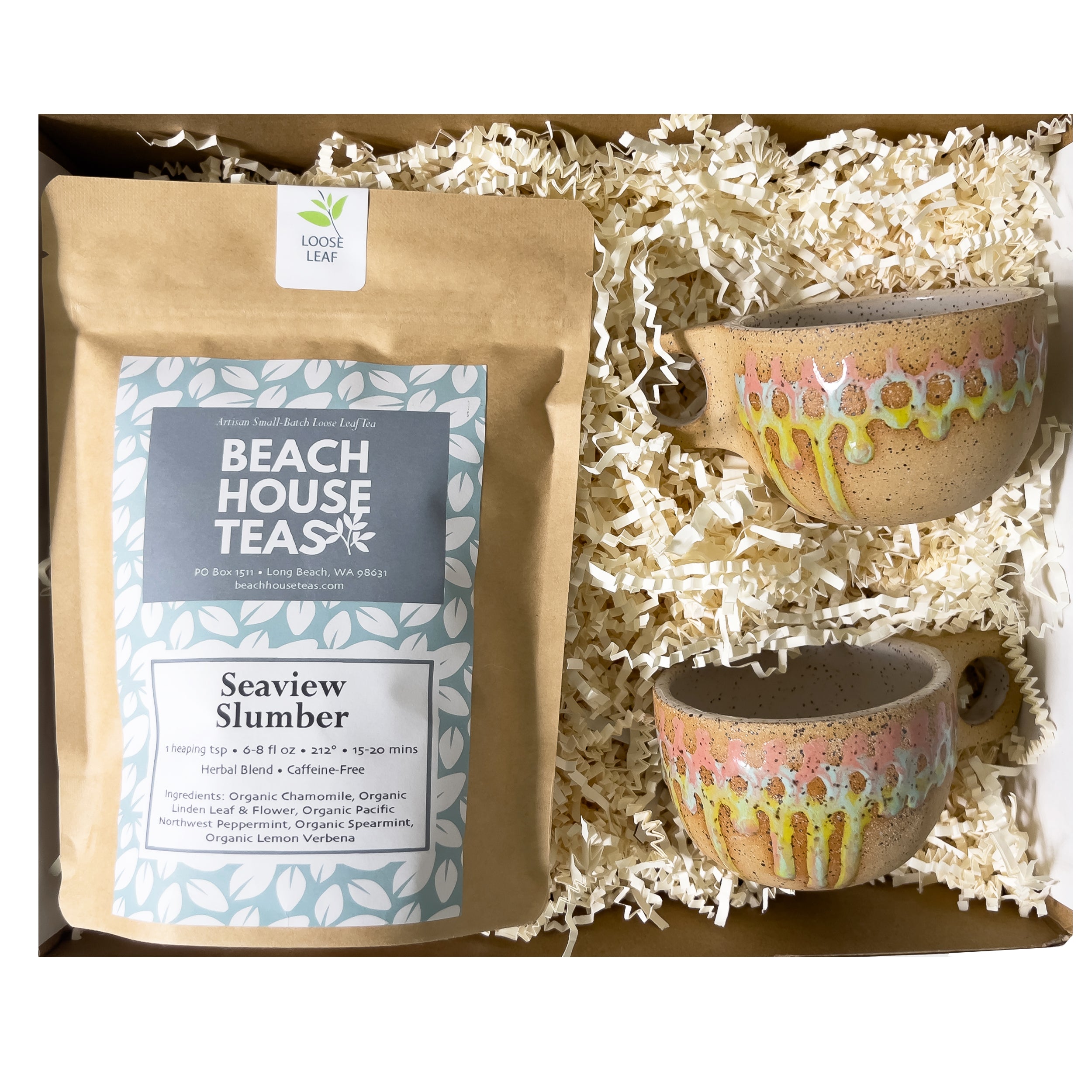 An overhead view of a bag of loose leaf tea called Seaview Slumber on the left and on the right two unique hand-crafted cappuccino style mugs stacked atop one another. All products are nestled in a box filled with shredded off-white crinkle paper. 